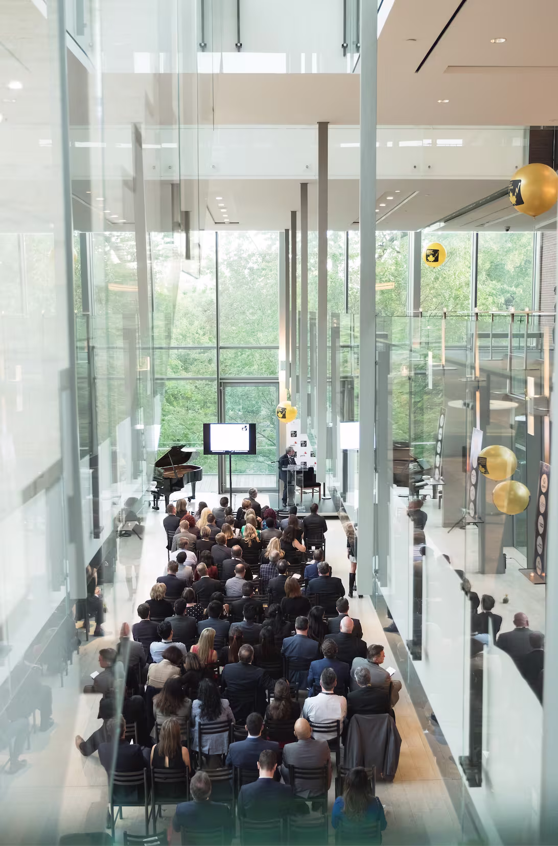 Speaker presenting to an audience in a bright glass atrium with a grand piano visible beside the stage.